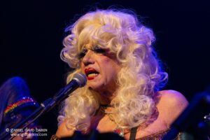 Performer with platinum blonde curls singing into a microphone on a dark stage, stage lighting shining on face