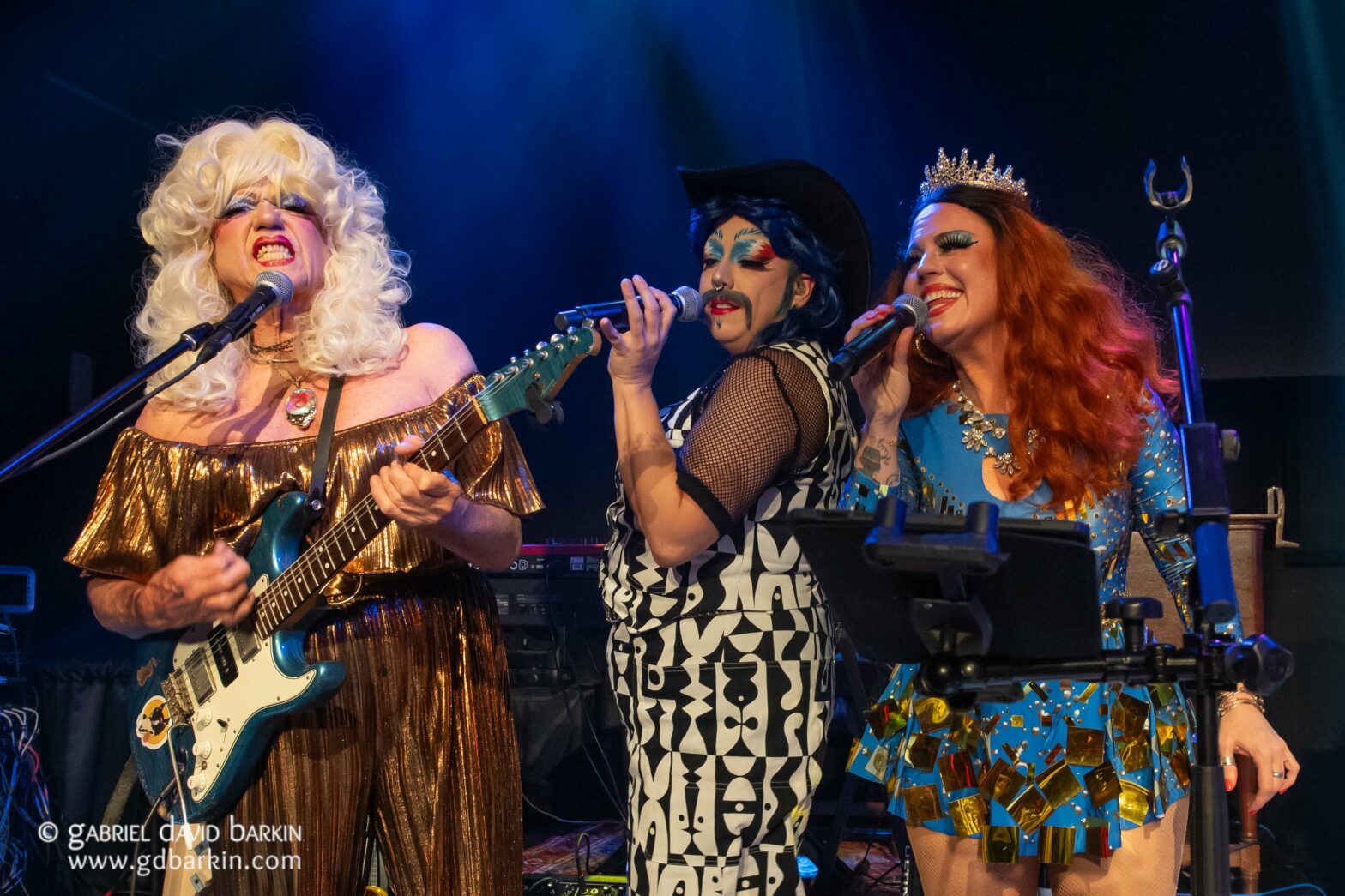 Three drag performers singing into microphones on stage, one with a white wig and teal guitar in a gold dress, the others in bold makeup and outfits with a tiara and blue sequins.