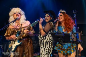 Three drag performers singing into microphones on stage, one with a white wig and teal guitar in a gold dress, the others in bold makeup and outfits with a tiara and blue sequins.