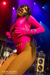 Live stage moment: performer in a bright pink bodysuit and fishnet tights dancing with hands on hips under red and blue stage lights, drums in the background