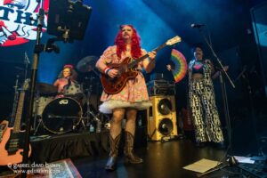 Lead guitarist with bright red wig in a pastel patterned dress plays a brown electric guitar on stage, with a drummer in the background and blue lighting above.