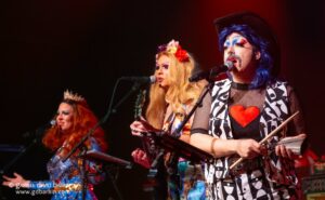 Group of performers in vibrant costumes and bold makeup performing on stage, with microphones and stage lighting visible. (One performer has blue hair and a heart motif outfit.)