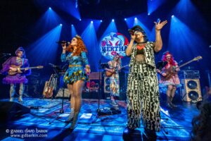 Drag performers on a brightly lit stage; a singer in a blue sequined dress at center, others with instruments, blue spotlights overhead and a circular backdrop logo behind them.