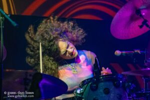 Drummer performing on stage under purple and pink lighting, with cymbals and drum kit visible.