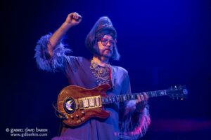 Performer playing a sunburst electric guitar on stage under blue lighting, wearing bold red glasses, ornate necklace, and feathered sleeves.