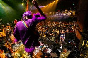 Front-of-stage view of a performer in a purple outfit and purple hair, raising a hand under colorful stage lights toward a cheering crowd.