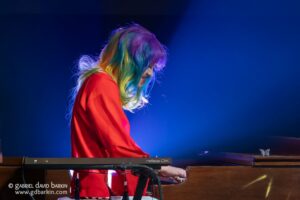 Musician with rainbow-colored hair playing a keyboard on stage against a blue backdrop, wearing a red outfit.