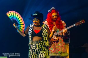 Two performers on stage: one in a black-and-white geometric outfit with a rainbow fan, the other with long red hair playing a bass guitar.