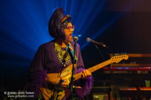 Person wearing a rainbow headband and red glasses sings into a microphone while playing an electric guitar on a blue-lit stage.