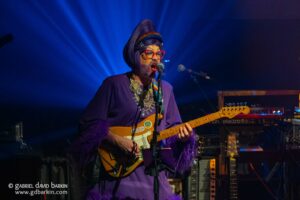 Performer in purple outfit with red glasses sings into a microphone while playing a Fender electric guitar on a blue-lit stage.