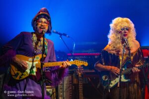 Two drag performers on stage with electric guitars and microphones under blue lighting.