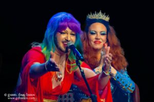 Two drag performers on stage singing into a microphone, one with a rainbow wig and red outfit, the other wearing a crown and blue outfit against a dark background
