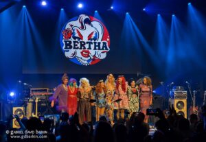 Group of performers in colorful costumes on stage with blue lights and a large Bertha logo on a screen behind them, audience in foreground taking photos