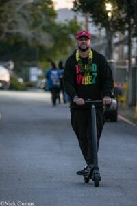 Man in a red cap and black hoodie rides a stand-up scooter down a city street, wearing a yellow-leaf lei around his neck.