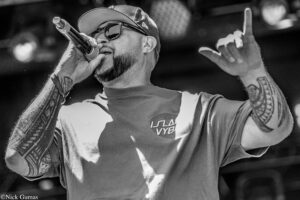 Male singer in a cap and sunglasses belts into a handheld mic, raising one finger on stage (black-and-white photo).