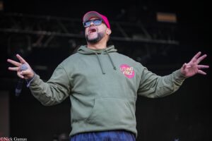 Male performer singing into a handheld mic with arms outstretched on stage, wearing a green hoodie and pink cap, dark background.