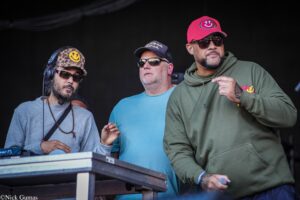Three men performing on a stage around a DJ booth: left in a camo hat with a yellow smiley patch and headphones, middle in a cap and sunglasses, right in a red cap and green hoodie holding a microphone.