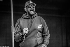 Man in a hoodie and cap, laughing with a microphone in hand on stage, wearing sunglasses.