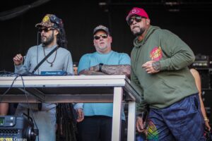 Three men wearing sunglasses stand behind a DJ booth on a dim stage, with cables and equipment visible in front of them.