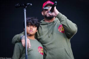 Adult man and boy on stage in matching olive hoodies; the man sings into a microphone while the boy rests his arm on his shoulder against a dark background.