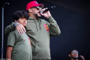 Man in an olive hoodie sings into a handheld microphone on stage, wearing a red cap and sunglasses, with a boy beside him.