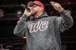 Male performer wearing a red cap and sunglasses, singing into a handheld microphone on stage, in a gray washed hoodie.