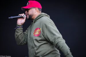 Male performer singing into a microphone on stage, wearing a red cap, sunglasses, and an olive-green hoodie with a circular patch on the chest.