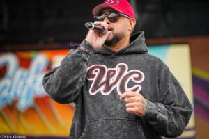 Male performer in a red cap and gray hoodie sings into a handheld microphone on a graffiti-covered stage.