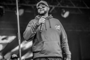 Male singer wearing a hoodie, cap, and sunglasses performs on stage with a handheld microphone.