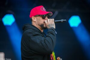 Male vocalist in a red cap and dark sunglasses singing into a microphone on stage, with a visible tattoo on his forearm.