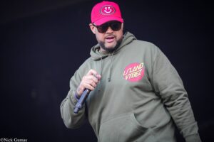 Stage performer wearing a bright pink cap with a circular logo, dark sunglasses, and an olive hoodie, holding a microphone while performing on stage.