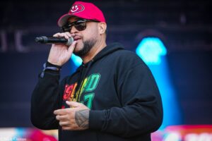 Male singer wearing a red cap and dark sunglasses, singing into a handheld microphone on stage? with blue lights in the background