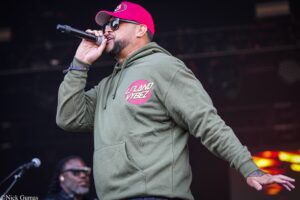 Male singer wearing a pink cap and sunglasses, singing into a handheld microphone on stage in a green hoodie with a pink circular logo.