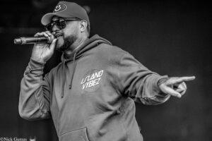 Male singer on stage wearing a hoodie and cap, holding a microphone and singing into the crowd (black and white photo).