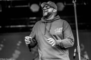 Male singer on stage wearing a hoodie and cap, holding a microphone and smiling, with stage equipment blurred in the background.