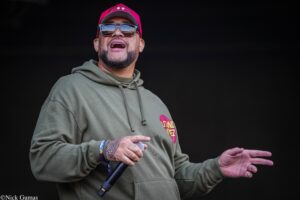 Male performer in an olive hoodie and pink cap, wearing sunglasses, holding a microphone and gesturing on stage from a low-angle shot.