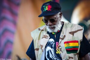 Portrait of an older man with a white beard wearing sunglasses, a camouflage vest, and a navy cap with a colorful embroidered lion. The background is blurred.