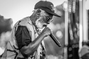 Black-and-white photo of a bearded man wearing a cap and sunglasses, singing into a handheld microphone on stage outdoors.