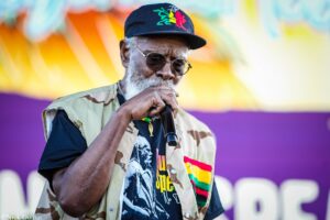 Older man wearing sunglasses and a camo vest sings into a microphone on stage, colorful mural behind him.