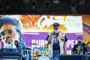 Live reggae band on stage performing, lead singer in camouflage vest and cap with a colorful banner behind him