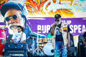 Male vocalist with sunglasses sings into a microphone on a colorful stage, graffiti-style mural behind him.