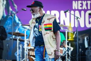 Older man with gray beard wearing a camouflage vest and black cap on a concert stage, holding a beaded bracelet; purple backdrop behind him.