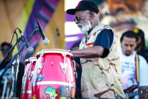 Older man with gray beard in a camouflage vest and cap playing red drums on a stage with microphones nearby.