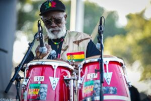 Older Black man with a white beard and sunglasses, wearing a cap and camouflage vest, playing red drums on an outdoor stage with microphones nearby.
