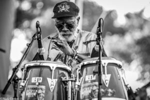 Older man with a white beard wearing sunglasses and a cap, playing a drum kit on stage.