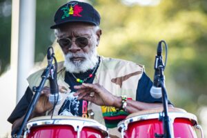 An older man with a gray beard wearing sunglasses and a camouflage vest, playing red drums outdoors with microphone stands nearby.