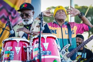 Two street musicians performing at an outdoor festival: a bearded man on red conga drums and a second man in a yellow knit cap playing a guitar down to his side.