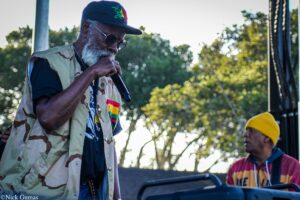 Older man in a camouflage jacket sings into a handheld microphone at an outdoor concert, wearing a dark cap and sunglasses with a bearded face visible behind the mic.