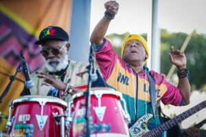 Two musicians performing outdoors: a guitarist wearing a yellow beanie and colorful jacket raises his fist, while a drummer with sunglasses plays nearby.