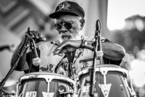 Close-up of an elderly man with a white beard wearing a cap and glasses, playing a drum kit with drumsticks in hand.